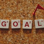 Scrabble tiles forming the word 'goal' with a small red flag on a cork board background.